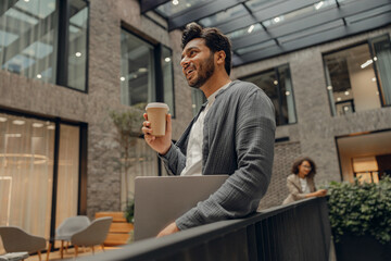Handsome businessman with laptop is standing on office background and drinking coffee