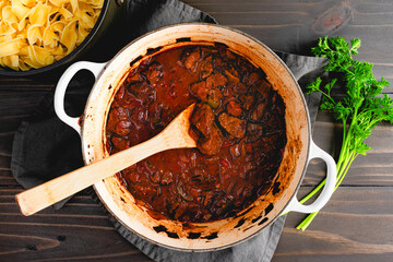 German Goulash Simmered in an Enameled Dutch Oven: Beef simmered in a seasoned tomato sauce in a cast-iron pot with a wooden spoon - obrazy, fototapety, plakaty