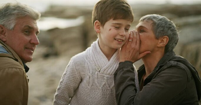 Secret, smile and grandparents at the beach with their grandchild for holiday or vacation together. Family, whisper and gossip with a senior woman speaking to a young boy while laughing outdoor