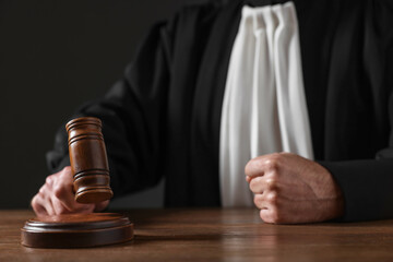 Judge with gavel sitting at wooden table against black background, closeup