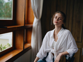 Woman sitting at home by a wooden window with a smile on a chair in homemade comfortable clothes and looking at the landscape, spring mood, women's day, rest on the weekend.