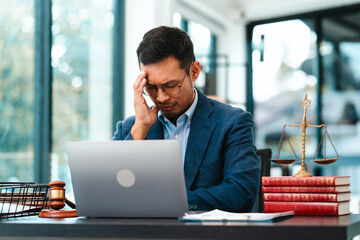 Asian male business lawyer looks stressed, pinching the bridge of his nose in front of a laptop at...