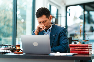 Asian male business lawyer looks stressed, pinching the bridge of his nose in front of a laptop at his office desk.