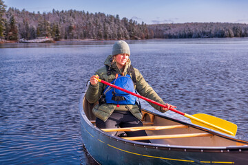young woman paddling a green canoe solo on a river with trees clad in freshly fallen snow in the...