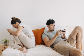 Asian Thai couple sitting back to back on sofa and using mobile phone, both man and woman living in apartment room.
