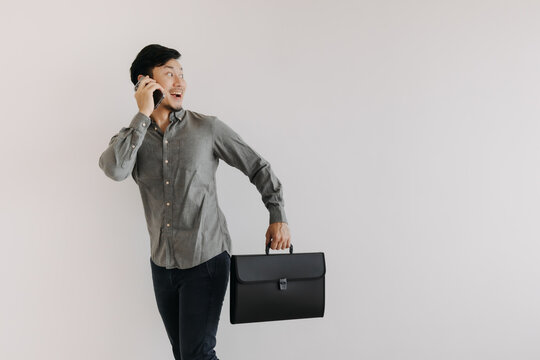 Happy Asian Thai Business Man Using Mobile Phone And Holding Black File Folder Bag While Walking Isolated Over White Background Wall And Looking At Empty Space..