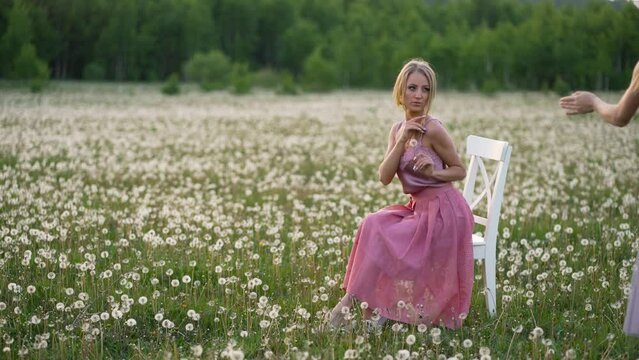 A woman in a skirt poses sitting on a white chair in a field for a photographer.