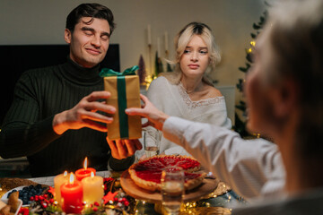Front view of happy surprised young man receiving Christmas gift from loving mother-in-law at home on xmas eve. Rear view of unrecognizable female giving present to handsome male on holidays.