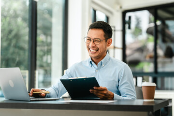 Middle-age Asian businessman smiles while looking at a laptop, possibly working as a Market Researcher or Marketing Analyst. - obrazy, fototapety, plakaty