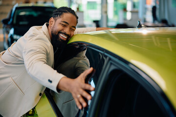 Happy African American man embracing his new car in showroom.