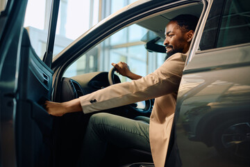 Happy African American man getting inside of his new car after buying them in showroom.
