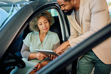 Black salesman and his female customer using touchpad at car dealership.