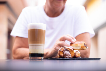 Unrecognizable elderly woman sitting at the cafe table in the morning having breakfast with sweet food and a glass cup full of coffee and milk.