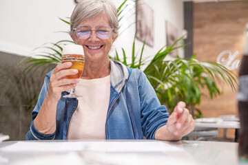 Smiling senior woman sitting at restaurant table with a glass of beer while choosing what to eat...