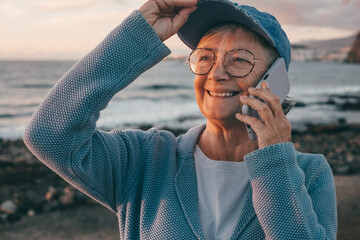 Handsome senior woman talking on phone standing at the sea beach at sunrise. Elderly smiling lady with cap and eyeglasses enjoying vacation or retirement © luciano