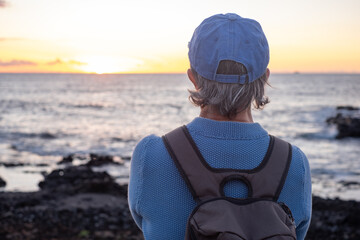 Back view of mature senior woman holding backpack looking at sunset at sea, horizon over water and...