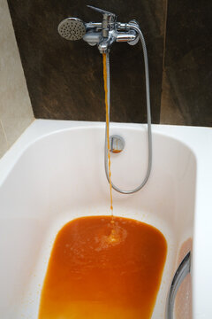 Rusty water flows from the faucet into the bathtub on the home bathroom. The flowing liquid from the tap in the domestic bath has turned a shade of red due to rust.