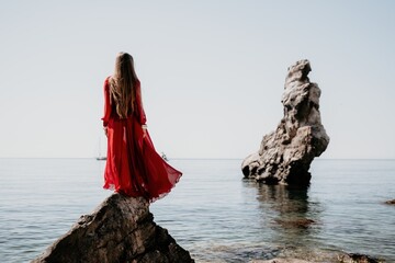 Woman travel sea. Young Happy woman in a long red dress posing on a beach near the sea on background of volcanic rocks, like in Iceland, sharing travel adventure journey