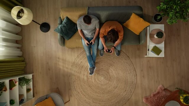 Top view shot of a homosexual couple at home. They are sitting on the couch, playing videogame with gamepads in their hands, having fun and play fighting.