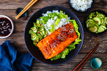 Fried teriyaki salmon steak with white rice and sliced cucumber on wooden table
