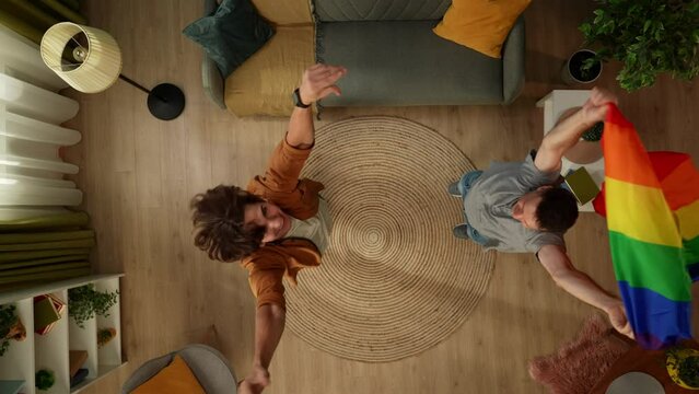 Top view shot of a homosexual, bisexual, pansexual, transgender couple jumping, dancing in the middle of the room, holding a LGBT flag to encourage love, equality, freedom.