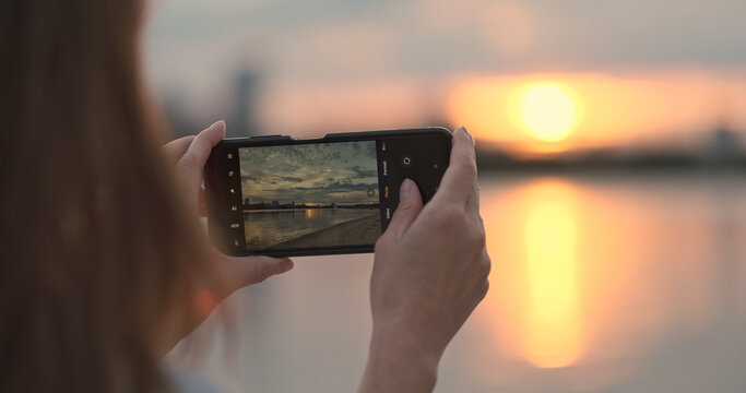Girl takes smartphone photo of sunset on wide river. Close-up. Orange sun reflects off surface water. Beautiful nature, impressions memory.