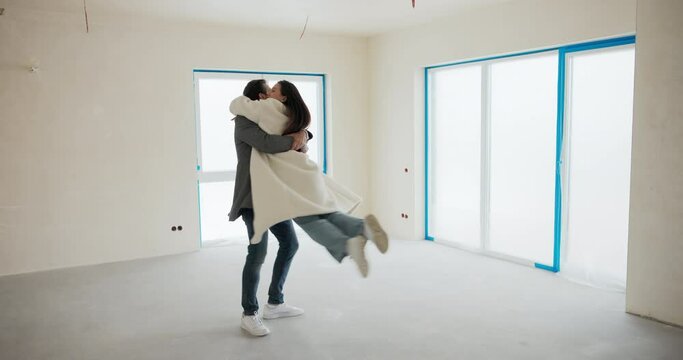 A couple in love stands in the middle of an empty apartment enjoying the purchase of a new home. The man in happiness takes his wife in his arms and spins around with her.