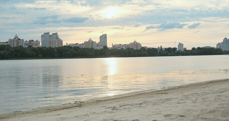 Sunset on beach in big city. Wide river, reflection sky water, sun's rays among clouds. High-rise residential buildings. Background