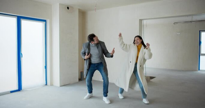 A young married couple warmly dressed dances playfully fools around in the middle of an empty room in their newly purchased home. A shell of a real estate investment.