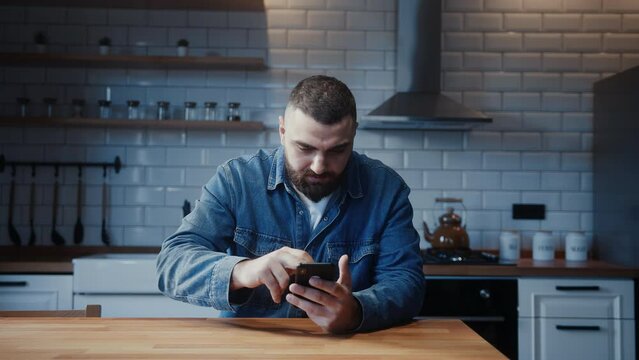 Young adult man sitting against the kitchen counter using smartphone having anxiety and stress. Upset man reacting to bad news