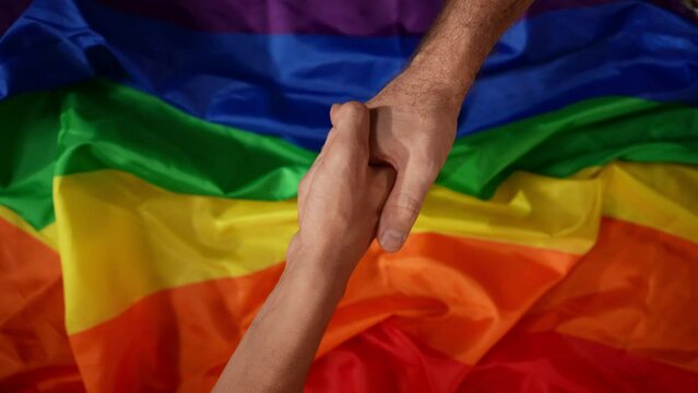 Top view detail shot capturing two male hands reaching for a handshake from the opposite corners of the frame. LGBT flag on the background.