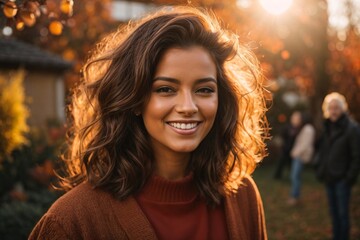 Smiling young brunette posing at a beautiful garden looking at the camera during late autumn sunset with a sun flares in the background, surrounded by friends and relatives.