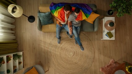 Top view shot of a homosexual couple at home. They are sitting on the couch wrapped in LGBTQ flag,...