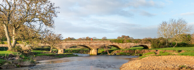 Panoramic picture of a stone bridge crossing a river in England. Beautiful landscape picture of rural England with soft clouds and blue sky behind.  