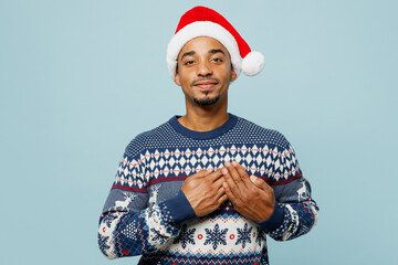 Young thankful grateful smiling man wear knitted sweater Santa hat posing put folded hands on heart...