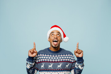 Young amazed man wear knitted sweater Santa hat posing point index finger overhead on area mockup...