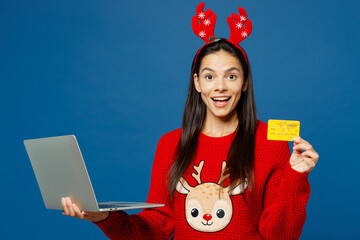 Merry young IT woman wears red Christmas sweater decorative fun deer horns on head posing hold...