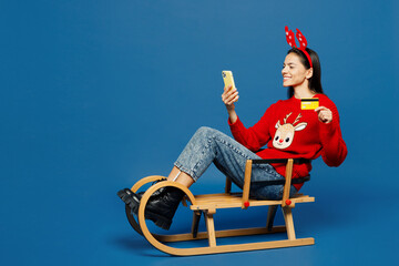Full body young Latin woman in red Christmas sweater decorative fun deer horns on head posing...