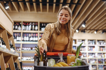 Young customer woman wear casual clothes put bottle of wine shopping at supermaket store grocery shop buying with trolley cart choose products inside hypermarket. Purchasing food gastronomy concept.