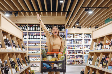Young fun customer woman wearing casual clothes do winner gesture shopping at supermaket store grocery shop with trolley cart buy choose food products inside hypermarket Purchasing gastronomy concept