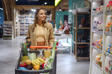 Young satisfied happy fun customer woman wear casual clothes shopping with trolley cart at supermaket store grocery shop buying choose products inside hypermarket. Purchasing food gastronomy concept.