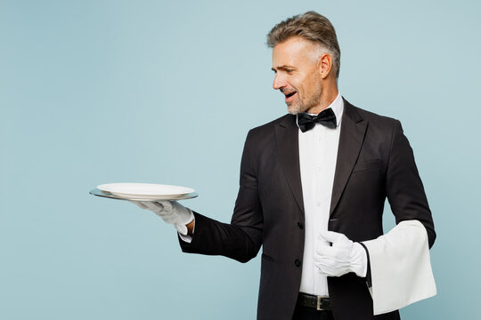 Adult sad barista male waiter butler man wear shirt black suit bow tie elegant uniform hold empty plate look camera towel work at cafe isolated on plain blue background. Restaurant employee concept.