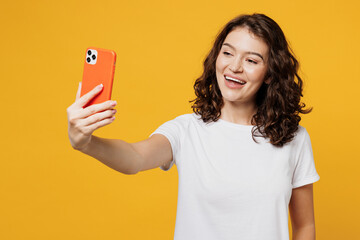 Young happy Caucasian woman she wear white blank t-shirt casual clothes doing selfie shot on mobile cell phone post photo on social network isolated on plain yellow orange background studio portrait.
