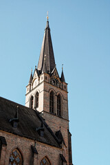 Obraz premium The image shows a brick church with a tall steeple and a clock on the tower. The church has arched windows and is lit by the sun against a blue sky. Spire on the bell tower of a Christian church.