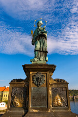 Obraz premium A statue of a saint holding a cross and a book on a pedestal. Statue of John of Nepomuk on Charles Bridge in Prague against the sky.