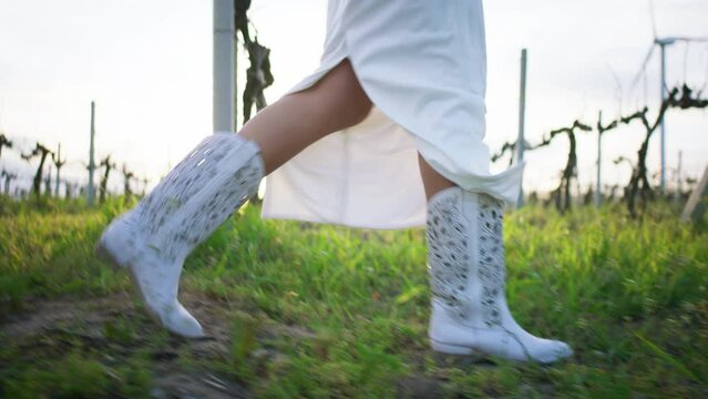 At sunset in the vineyard side walking in front of the camera lady wearing white cowboy boots and white dress
