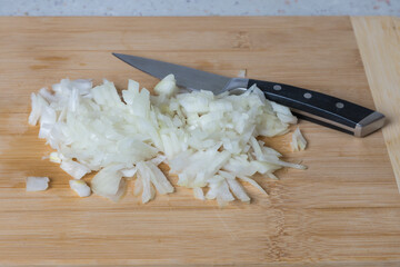 Finely chopped white onion. A knife and onion lie on a wooden cutting board.