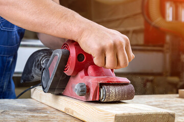 Young man builder carpenter equals polishes wooden board with a  random orbit sander  in the...