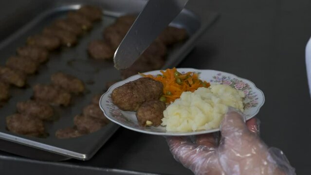 A female cook in a white coat puts a cutlet in a plate of potatoes in a restaurant kitchen.