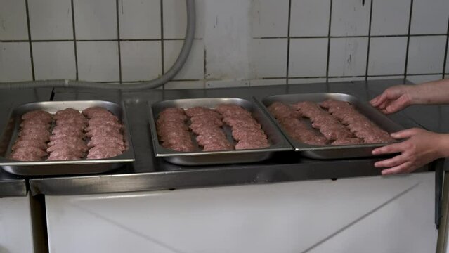 A female cook in a white coat lays a tray of raw cutlets on a table in a restaurant kitchen.